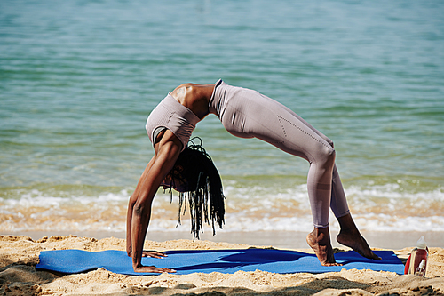 Young slim flexible Black woman doing wheel pose on the beach