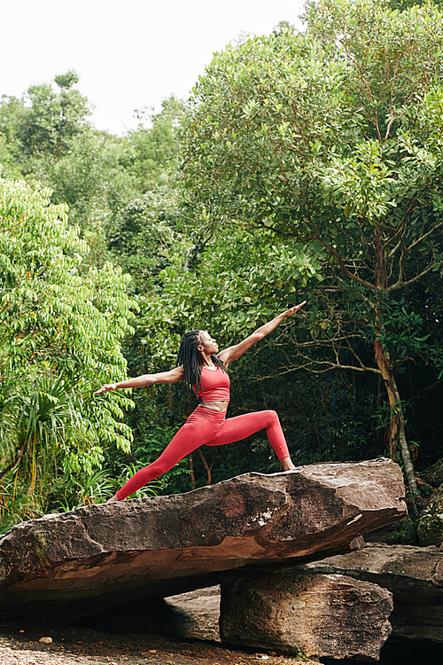 Young slim Black sportswoman enjoying practicing yoga on big rock in forest