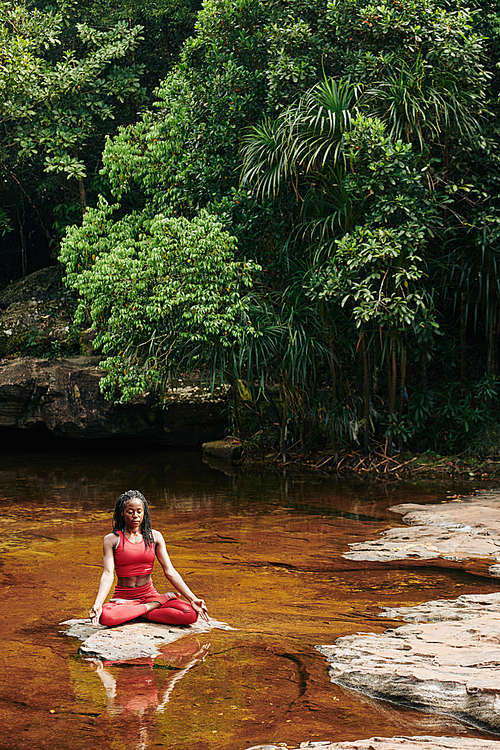 Pretty young Black woman sitting on rock in the creek in the center of forest and meditating