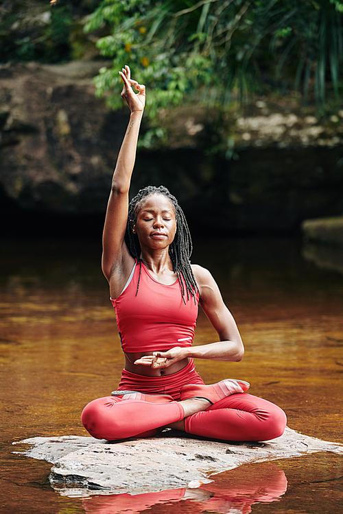 Concentrated young Black woman doing gyan mudra when sitting on rock in lotus position