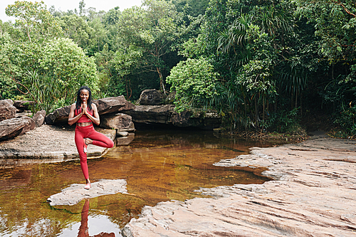 Positive woman standing on rock in creek in the center of wild forest and doing namaste gesture when standing in tree position