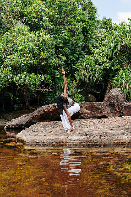 Sportswoman in cotton pants bending knees and looking at her raised arm when exercising in forest by water