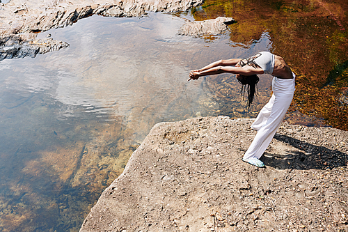 Black young woman in sportswear doing back bend when standing on rock by the river