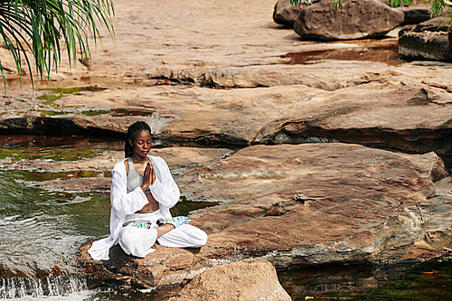 Beautiful calm young Black woman sitting on rock by the creek holding hands in namste gesture and meditating
