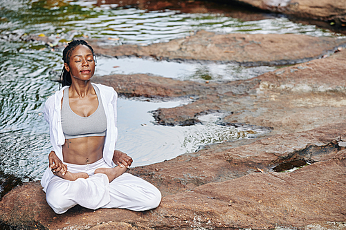 Serene young fit Black woman sitting in lotus position by the river, enjoying fresh air and legs stretch