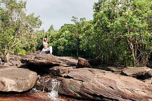Young pretty Black woman doing sun salutation flow when standing on rock by small waterfall in forest