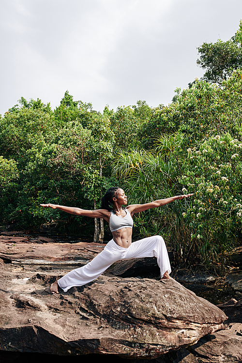 Beautiful young Black woman standing on big rock in remote forest in warrior two position and enjoying outdoor yoga session