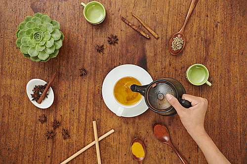 Close-up of woman pouring green tea from teapot into her cup while sitting at wooden table with different spices and seasonings