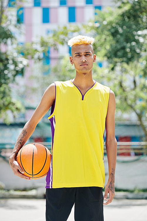 Young serious athlete with fashionable hairstyle holding basketball ball and 
