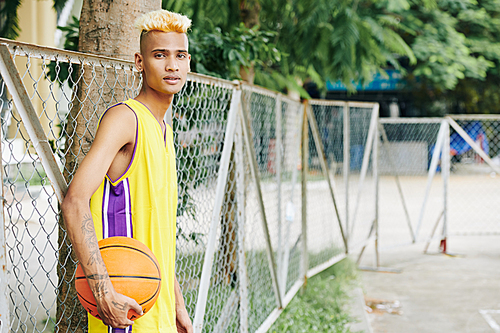 Handsome young slim player leaning on metal net around street basketball court and 
