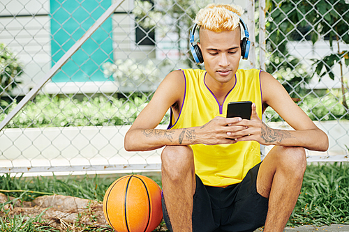 Young handsome man sitting on street basketball court and listening to music in smartphone application via headphones