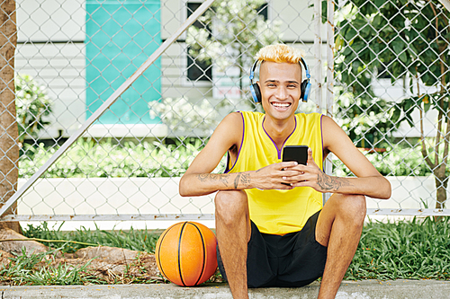 Portrait of happy laughing young Black basketball player sitting outdoors with smartphone in hands and enjoying music in headphones