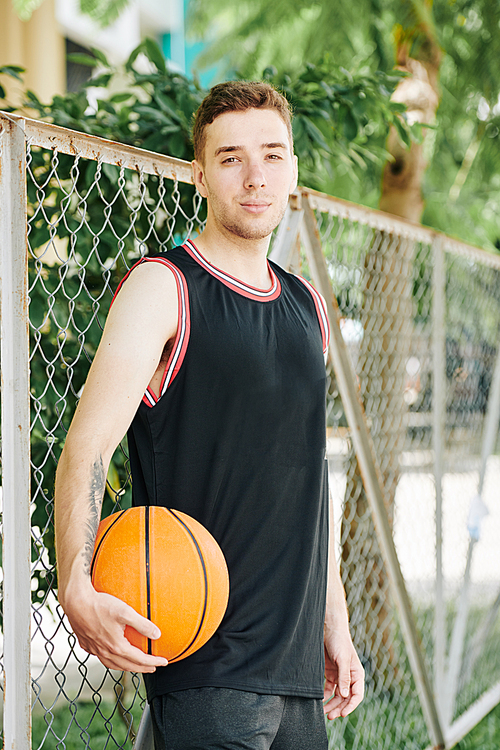 Positive handsome young man in black sports uniform standing at net around outdoor basketball court and 