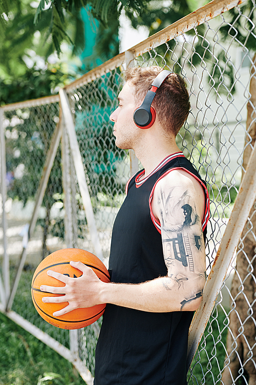 Handsome sporty young man leaning on net around street basketball court and listening to music in headphones when having break between quarters