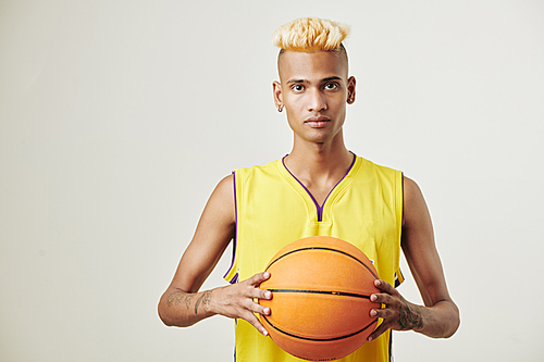 Portrait of serious young sportsman in yellow uniform posing in studio with baskball ball