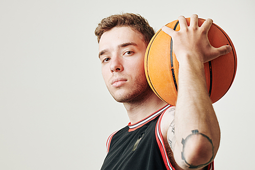Studio portrait of young serious sportsman holding basketball ball on his shoulder