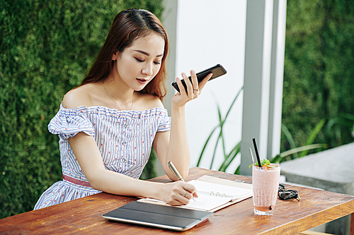 Young Vietnamese woman listening to voice message on her smartphone and taking notes in planner