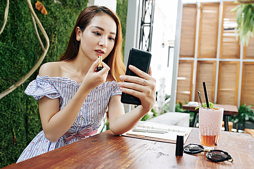 Beautiful young Asian woman sitting at cafe and using smartphone screen as mirror when applying red lipstick
