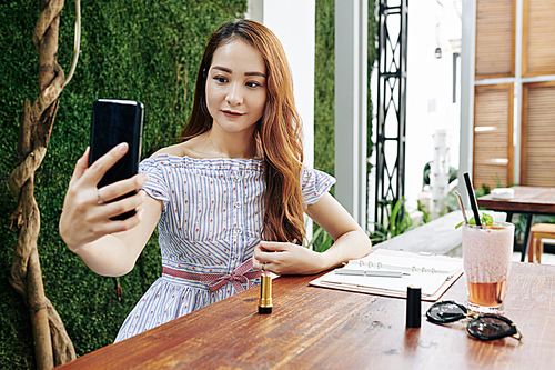 Pretty young Asian woman taking selfie on smartphone after applying new brown lipstick