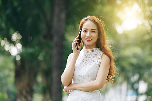 Portrait of young beautiful Vietnamese woman with long hair standing outdoors and talking on phone