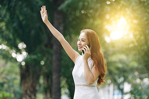 Attractive young Vietnamese woman talking on phone and waving with her hand when trying to catch taxi