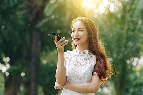 Young beautiful Vietnamese woman in white dress standing in park on sunny day and recording voice message