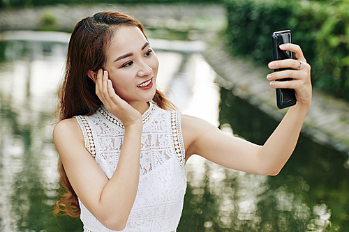 Young beautiful Asian woman posing for photo on smartphone when standing at pond in city park