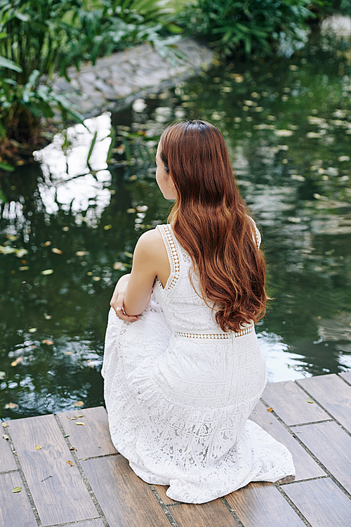 Young woman with long hair sitting at small pond in park and enjoying beauty of nature, view from the back