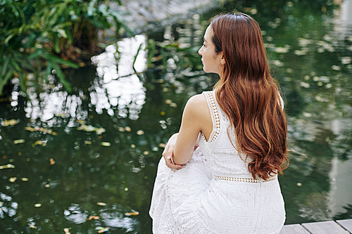 Dreamy young woman resting at small pond and looking at water, view form the back