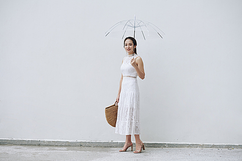 Young pretty Vietnamese woman in white lace dress standing outdoors with transparent umbrella