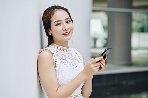 Portrait of young beautiful Asian woman with smartphone in hands leaning on the wall and 