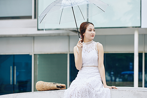 Charming young Vietnamese woman in lace dress sitting outdoors with transparent umbrella and looking away