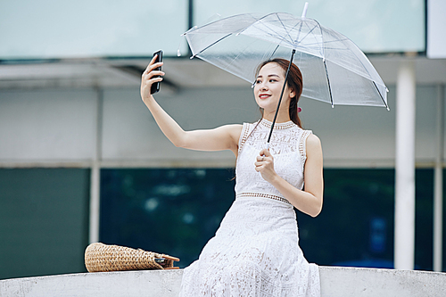 Attractive smiling young Vietnamese woman holding umbrella when taking selfie in city street