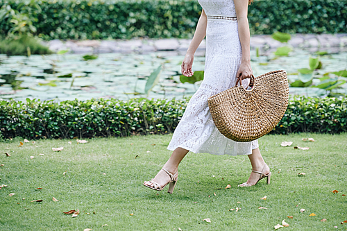 Cropped image of young woman in lace dress walking in park with straw bag on summer day