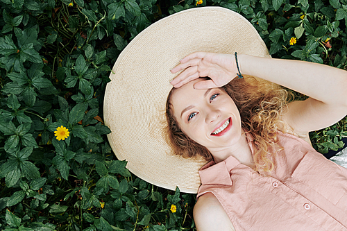 Happy young Caucasian woman in big straw hat lying on grass in park and lookig at camera, view from above