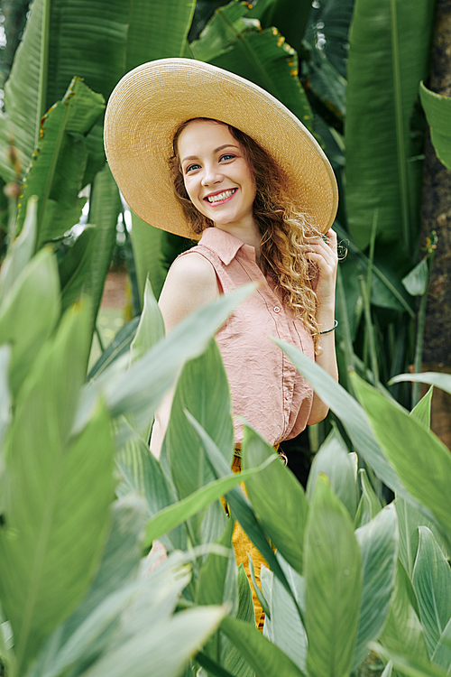 Beautiful young woman in big straw hat enjoying walking among flowers in summer garden