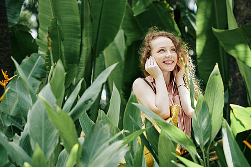 Happy young woman standing in tropicsl garden and listening to music in earbuds