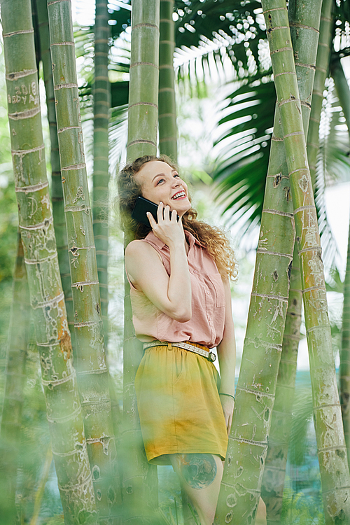 Happy young woman leaning on bamboo tree and talking on phone with friend