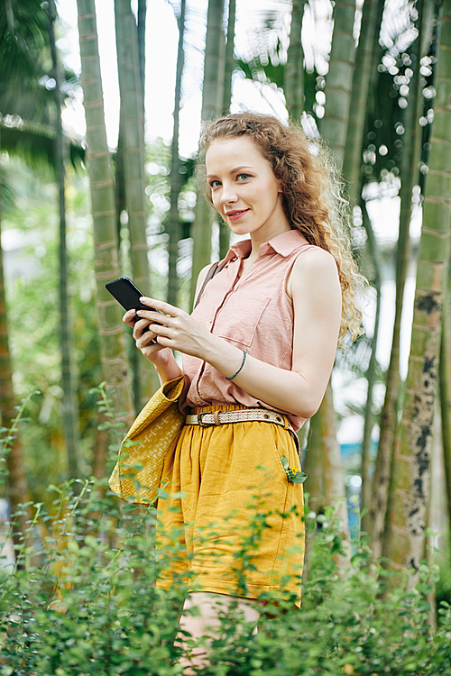 Portrait of petty young smiling woman in casual clothes walking in park and texting friends