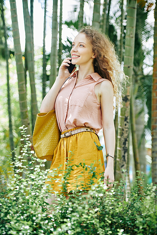 Portrait of happy inspired young woman standing in summer park and talking on phone with friend