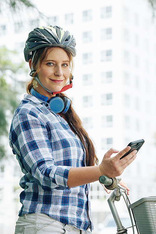 Portrait of happy young woman in bicycle helmet standing outdoors with her bicycle and sending text messages to friends