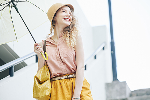 Attractive happy young woman in hat walking outdoors with umbrella on rainy day
