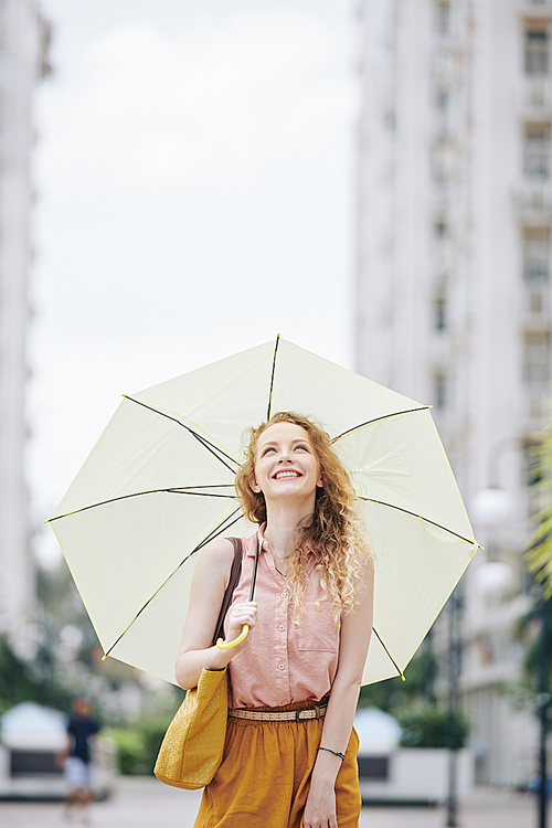 Happy emotional young woman with blond curly hair walking in the street with big umbrella and looking in the sky