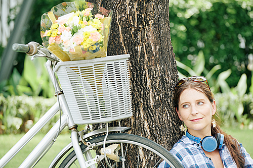 Dreamy smiling young woman sitting under the tree in park and looking at flower bouquet in basket of her bicycle