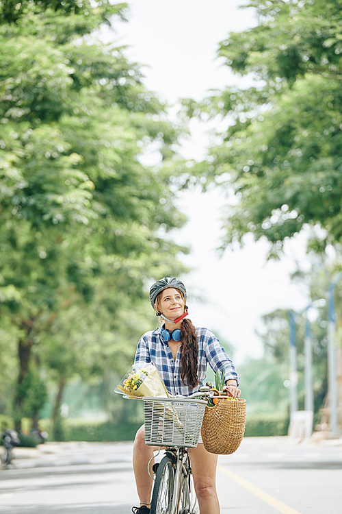Smiling happy young woman with flower bouquet and basket of groceries riding bicycle in city park