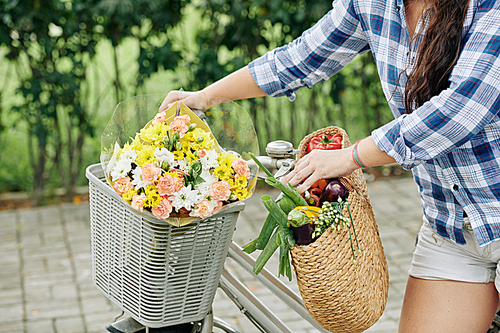 Young woman with basket of groceries and bouquet of flowers standing at her bicycle