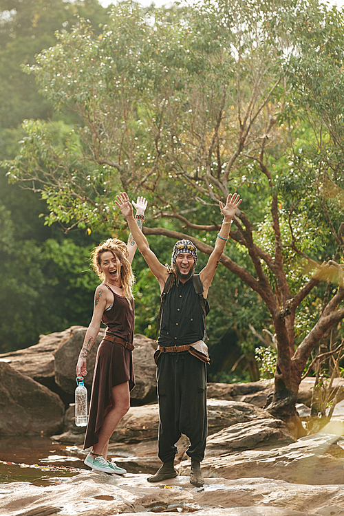 Happy excited middle-aged couple with dreadlocks rising hands when standing at brook with fresh water they can drink during trip in jungles