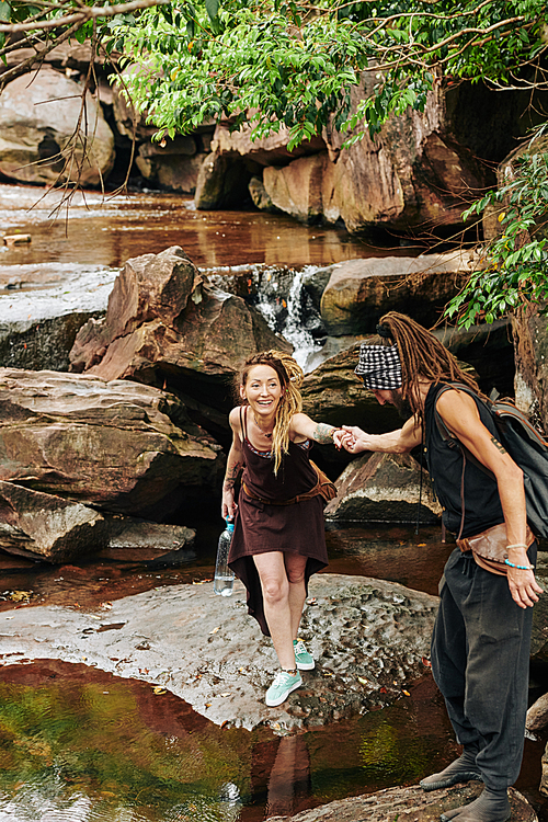Man with dreadlock giving hand to his beautiful smiling girlfriend and helping her to overstep the stream