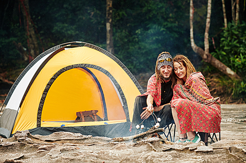 Happy mature couple with dreadlocks wrapping in blanket when sitting by fire near tent and frying marshmallows