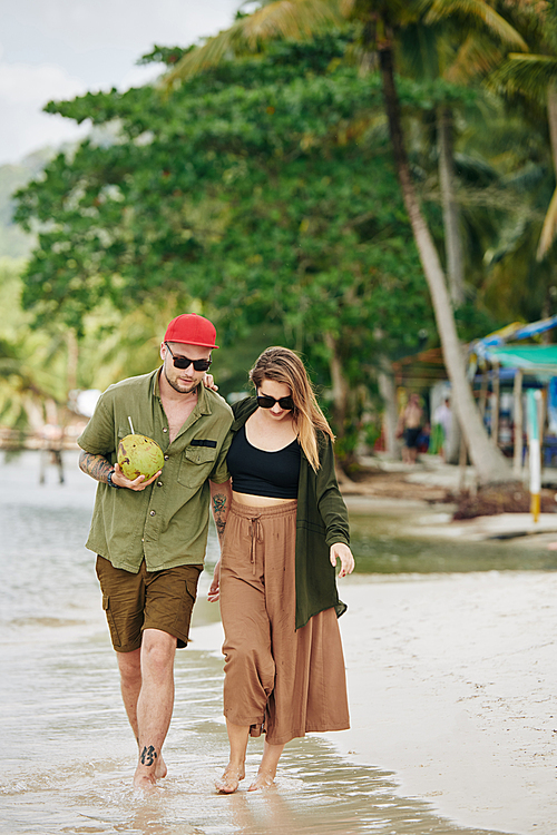Young couple in casual clothes walking on the beach and drinking coconut cocktail